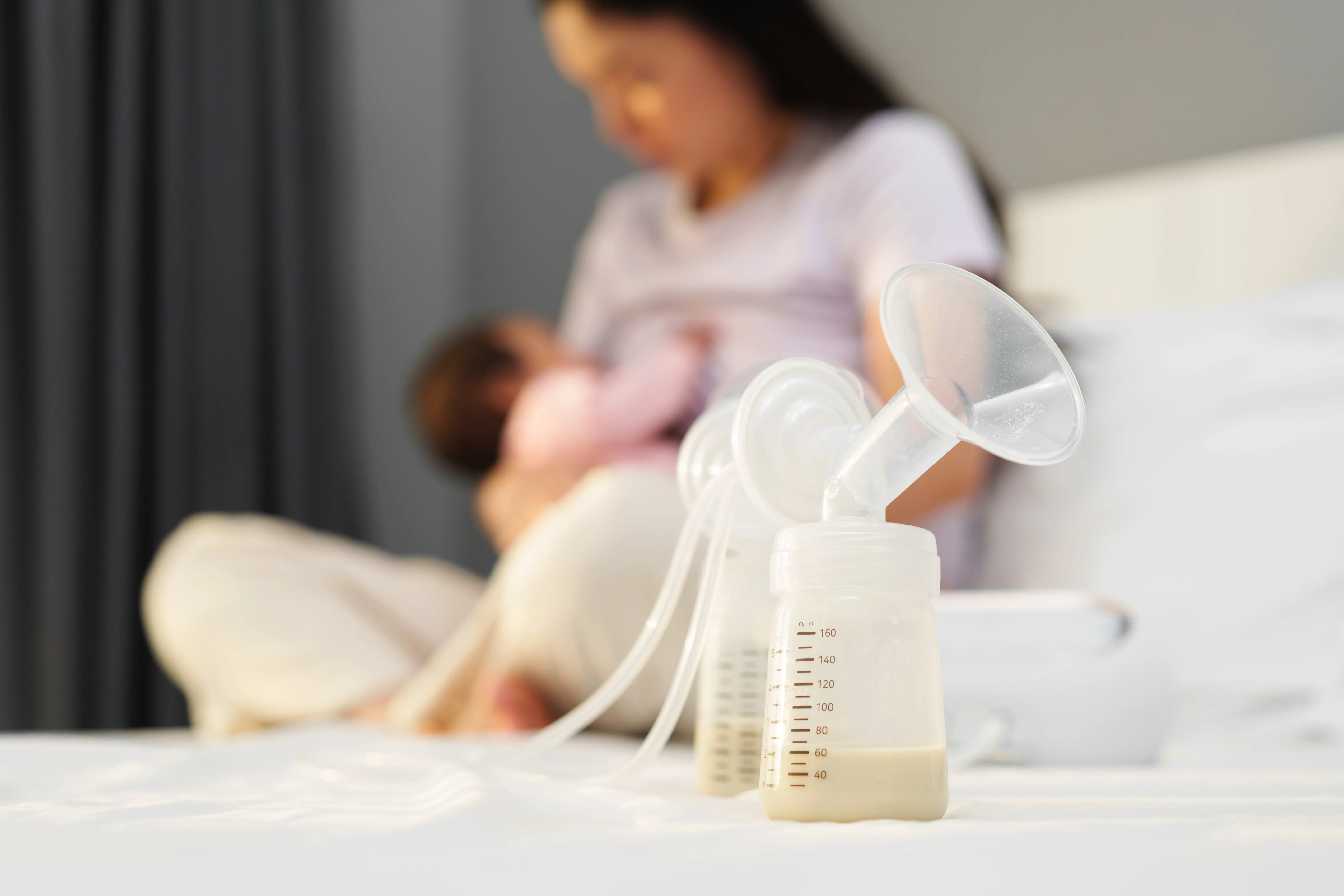 Breast pump and bottles in the foreground, mother holding baby in a blurred background
